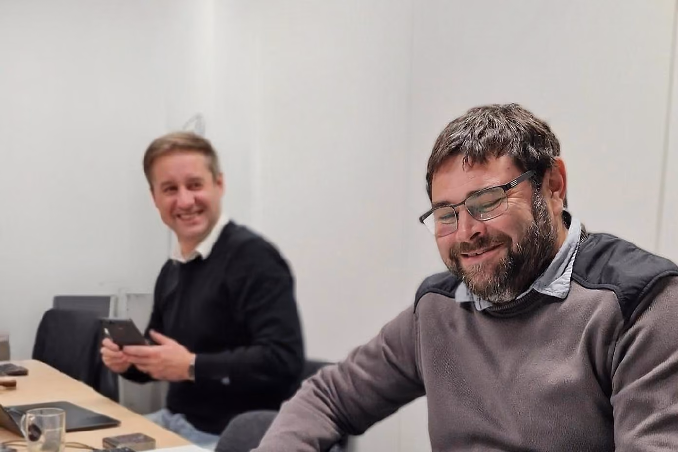 Two men sitting at a table in an office, one smiling holding a phone, the other smiling with glasses.