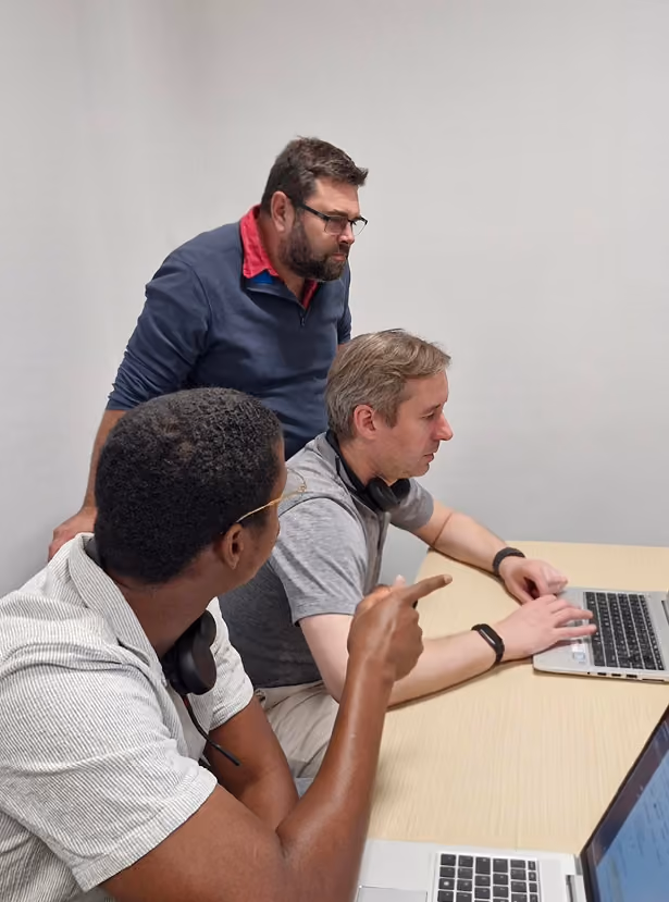Three men working together at a table with laptops, one man pointing at the screen.