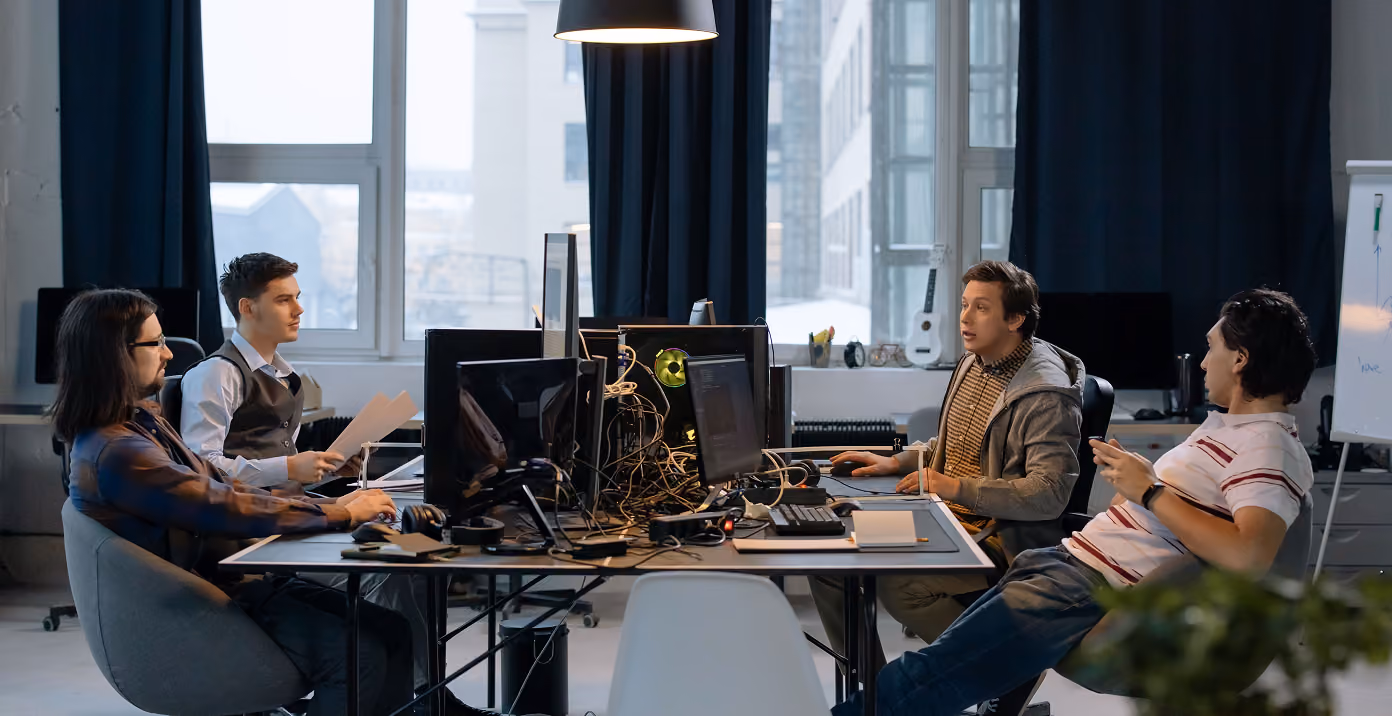 Four men sitting at a table working on computers and discussing in an office with large windows.