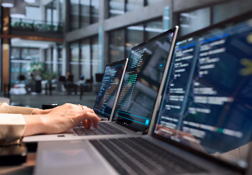 Hands typing on a laptop keyboard with multiple laptops displaying code in a modern office.