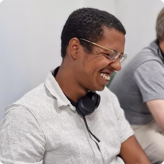 Smiling man wearing glasses, white shirt and black headphones around neck, seated indoors.