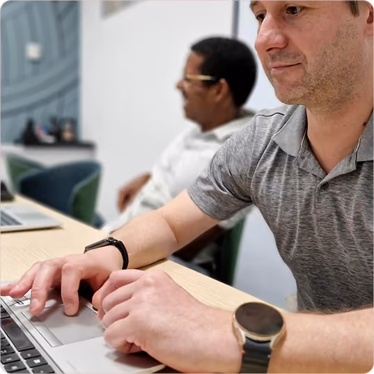 Man in gray shirt using laptop, another man blurred in background in office setting.