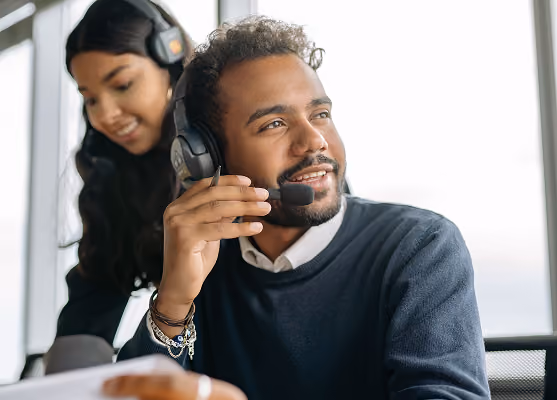Man wearing headset, smiling and looking away while woman with headset stands behind him.