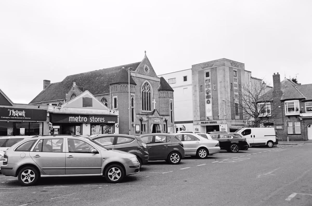 A black and white photo of Marlborough Square in Coalville, England, featuring a bingo hall, an off licence, an Asian restaurant and a church, with cars parked outside.