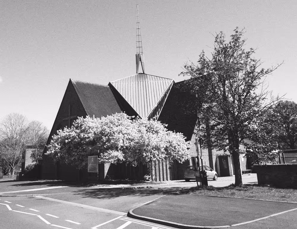 A black and white photo of the Church of the Epiphany in Corby, England, featuring a steeple and blossom trees on the surrounding road.