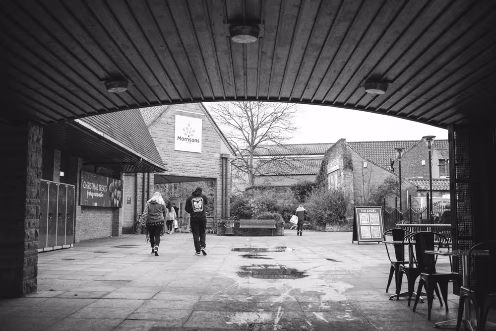 A black and white photo of the Woodhouse Centre shopping area in Mansfield, from under an archway, wit Morrisons supermarket and pedestrians visible.