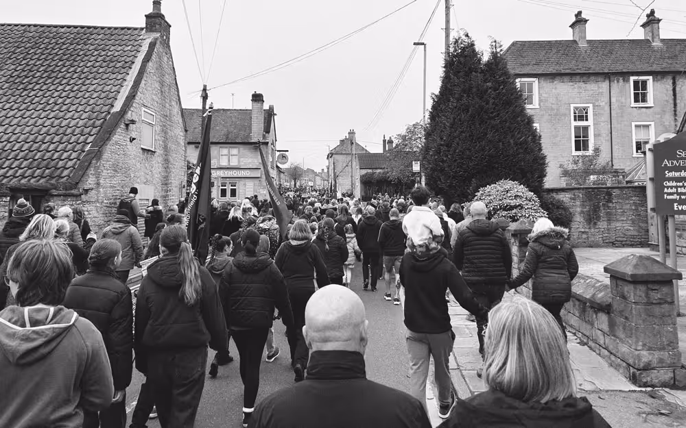 A black and white photo of Mansfield Remembrance Day in 2025, with crowds of people walking down a residential street, holding old folded up banners.