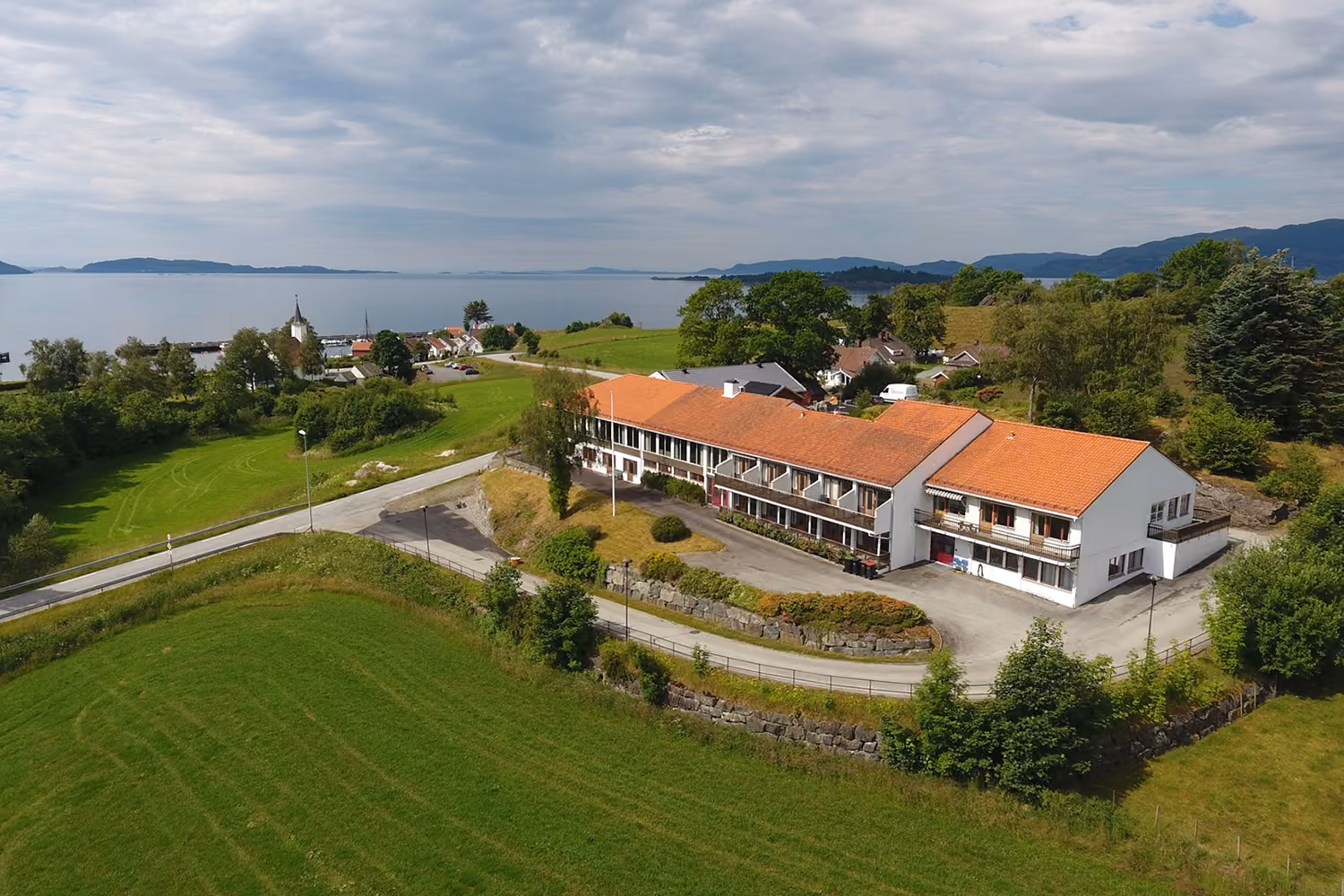 Flyfoto av et hotellbygg med røde tak i et grønt, kupert landskap nær sjøen.