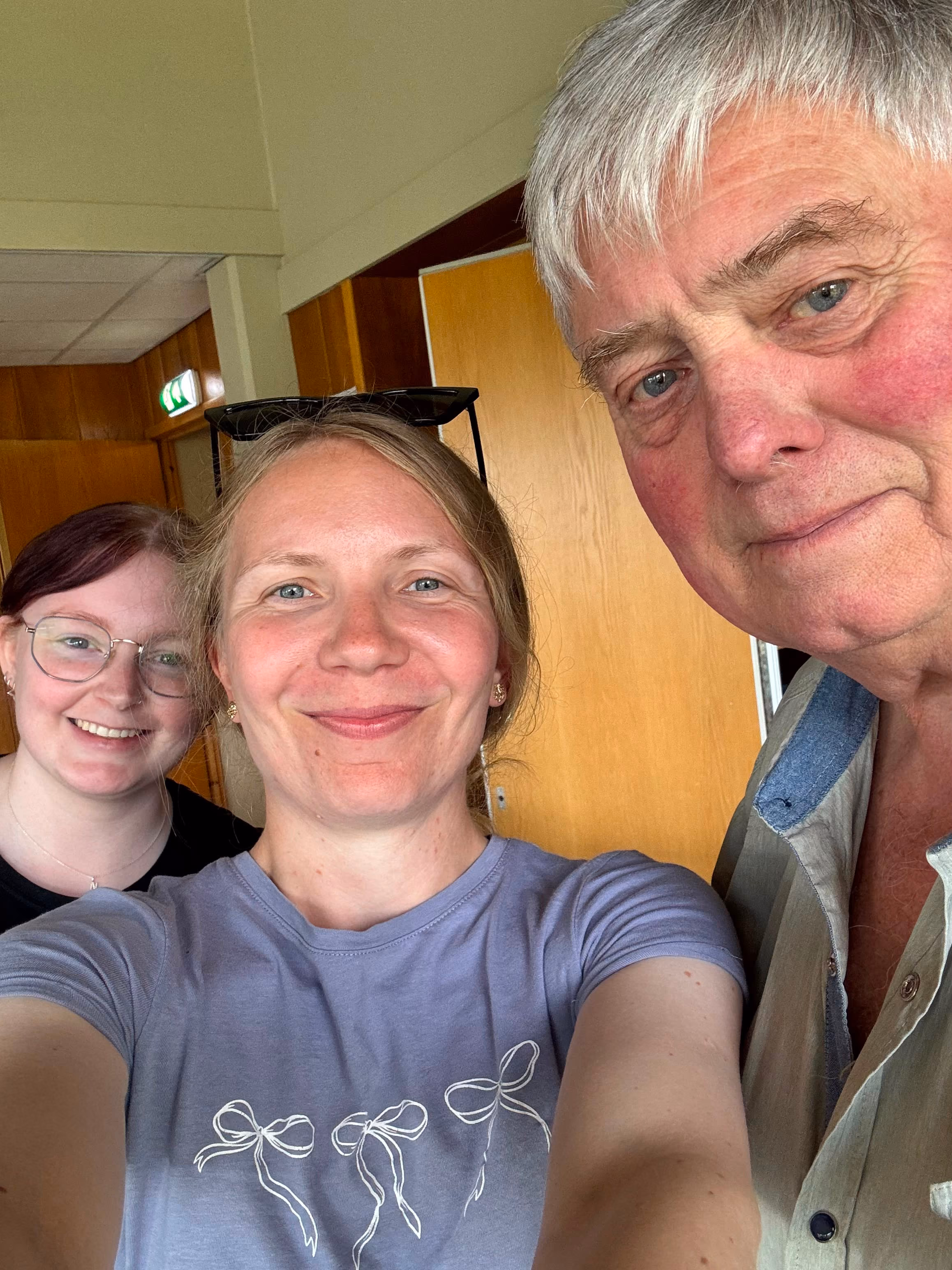 To kvinner og ein eldre mann som smiler og tek selfie inne i ei trepanelert rom.