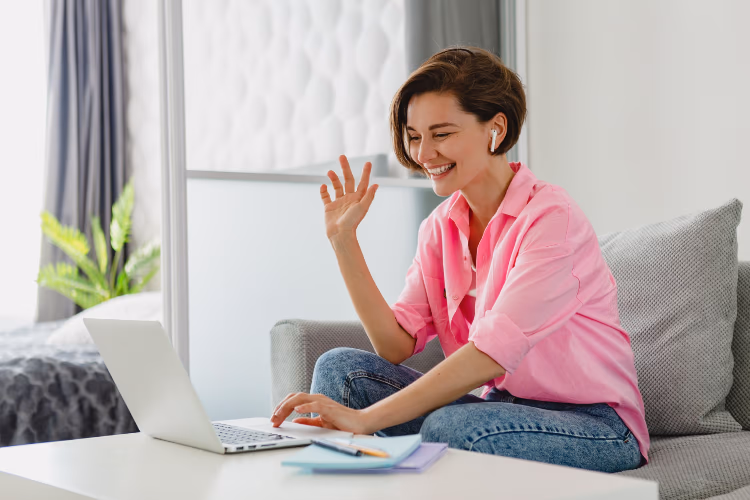 Woman on work call wearing earbuds, smiling and waving at laptop