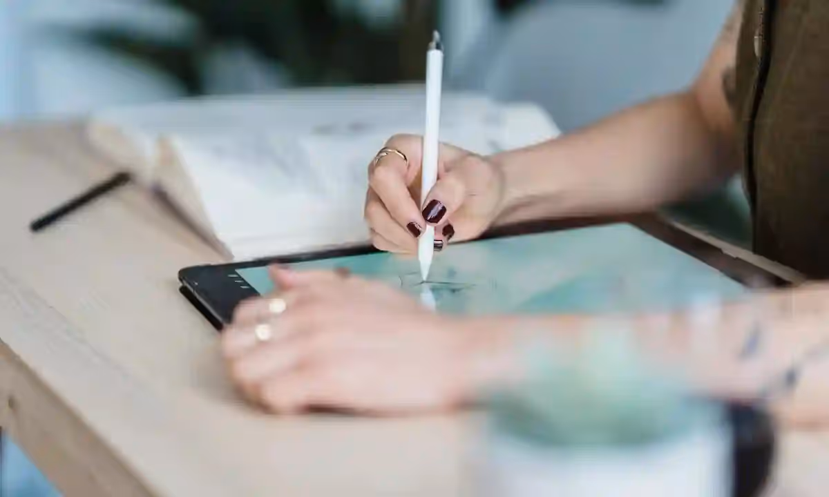 Crop unrecognizable female designer using tablet and stylus for drawing sketches on wooden table