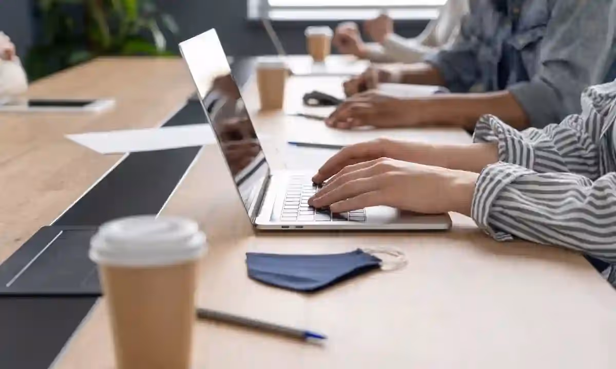 A businesswoman typing on a laptop at a bright indoor workspace with notebooks and a pen.