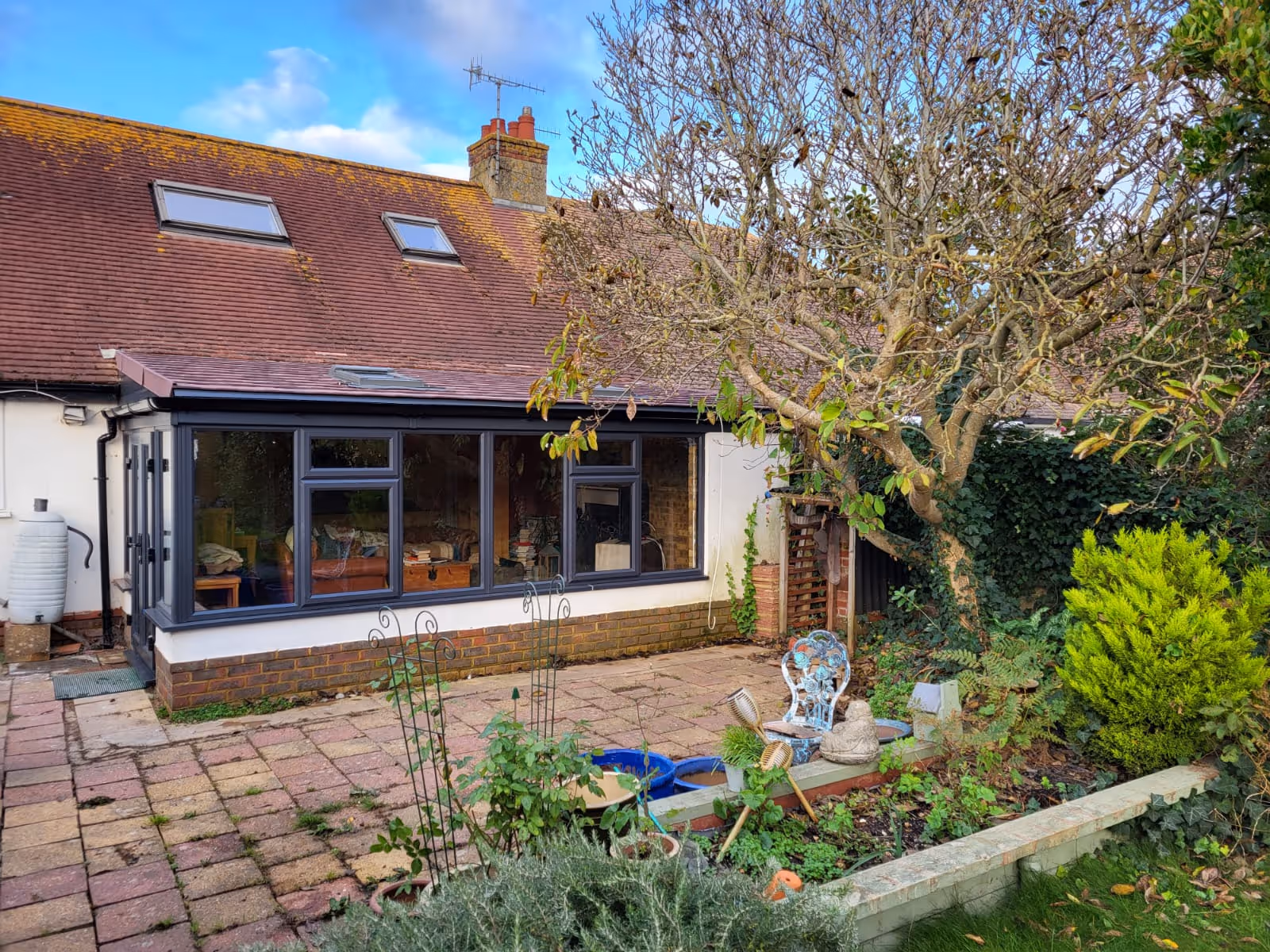 A house with large windows overlooks a patio garden with a tree and shrubs.