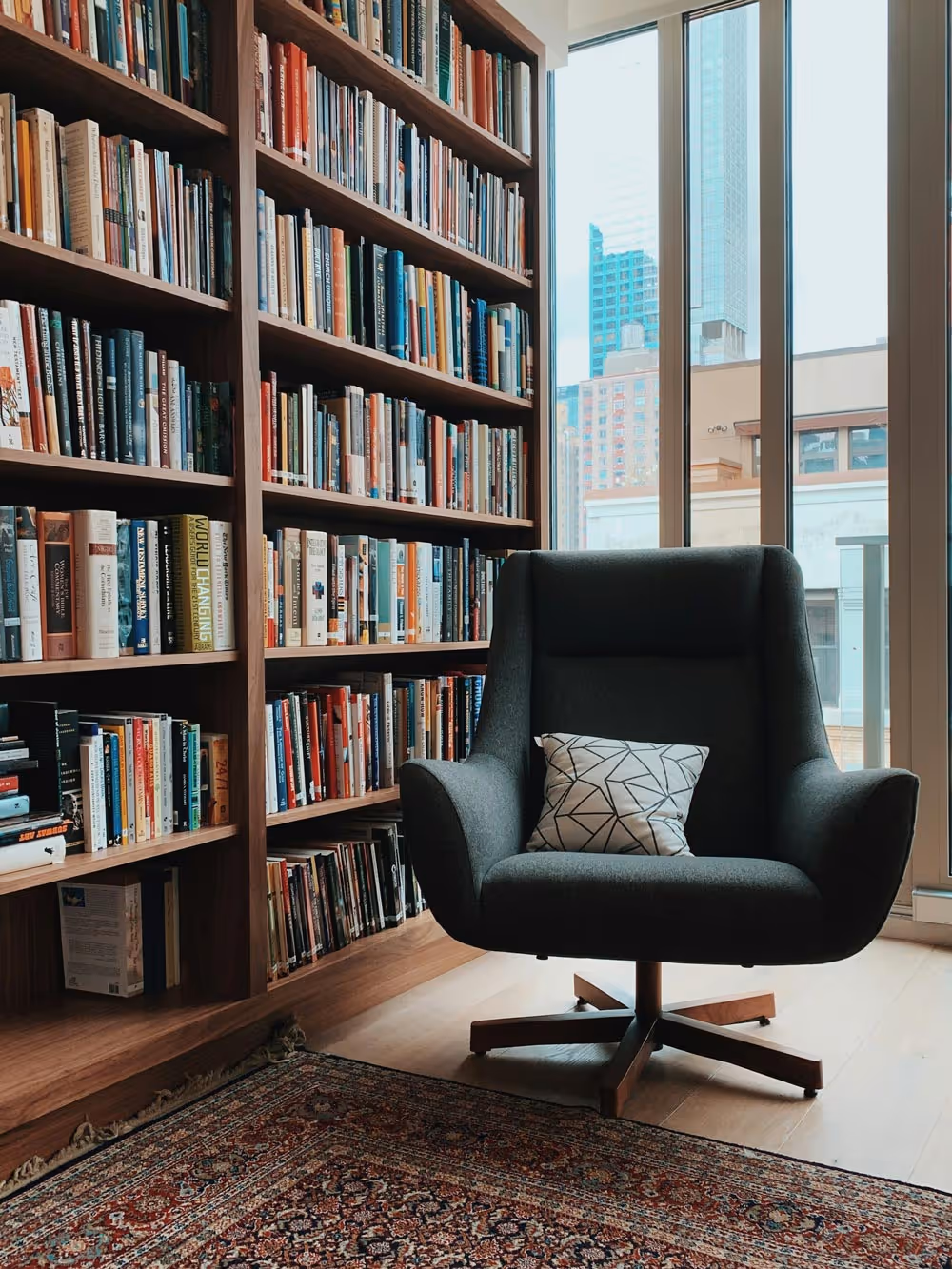 A cozy reading corner with a dark armchair, patterned pillow, and a bookshelf filled with books.