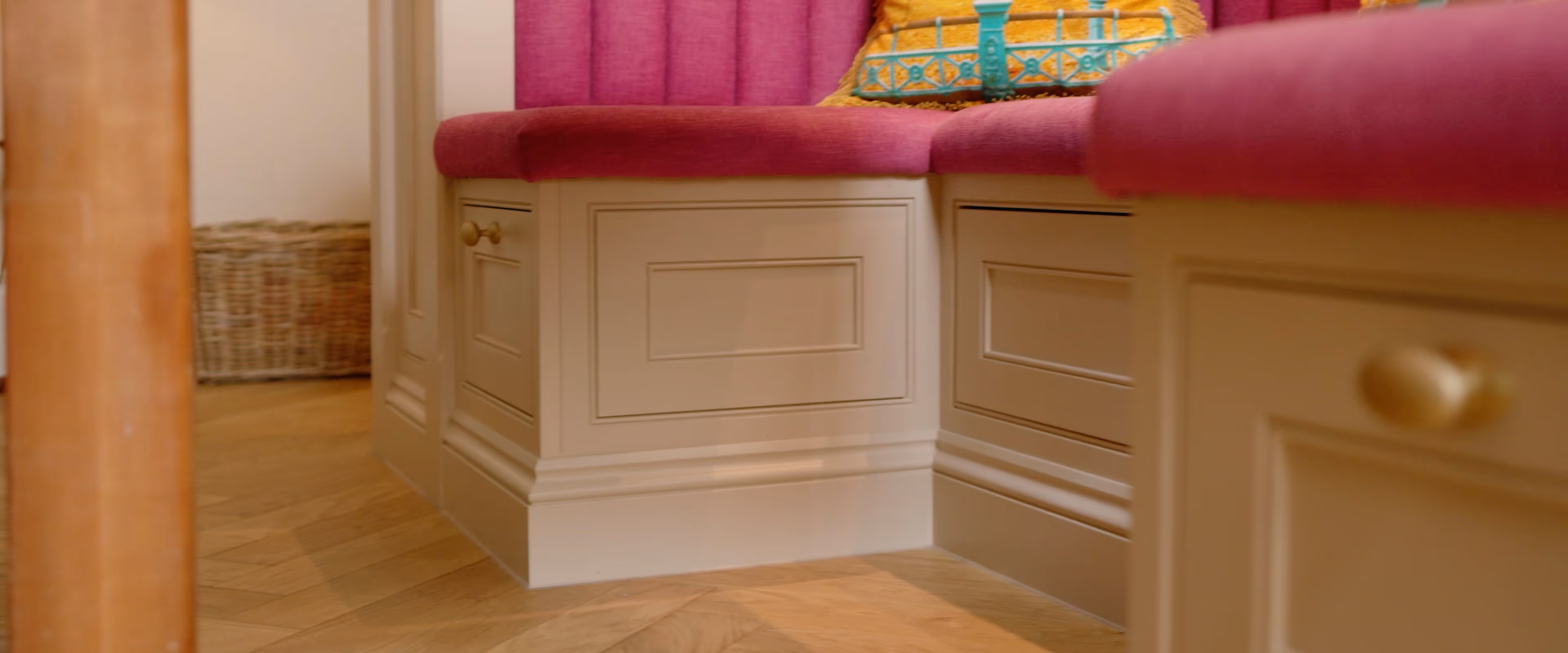 A cushioned corner bench with magenta upholstery and colorful pillows, against beige paneled cabinets.