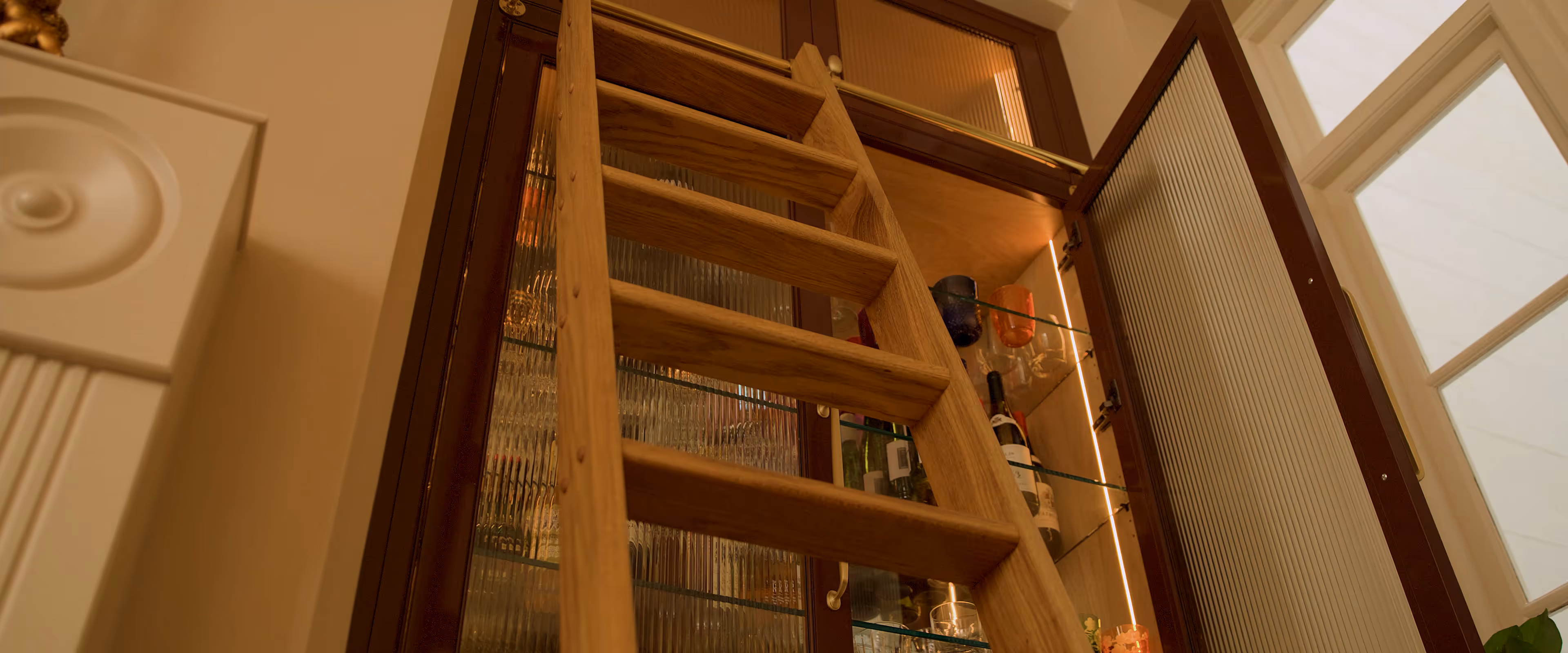 A wooden ladder is leaning against an open cabinet with shelves holding bottles and glassware.