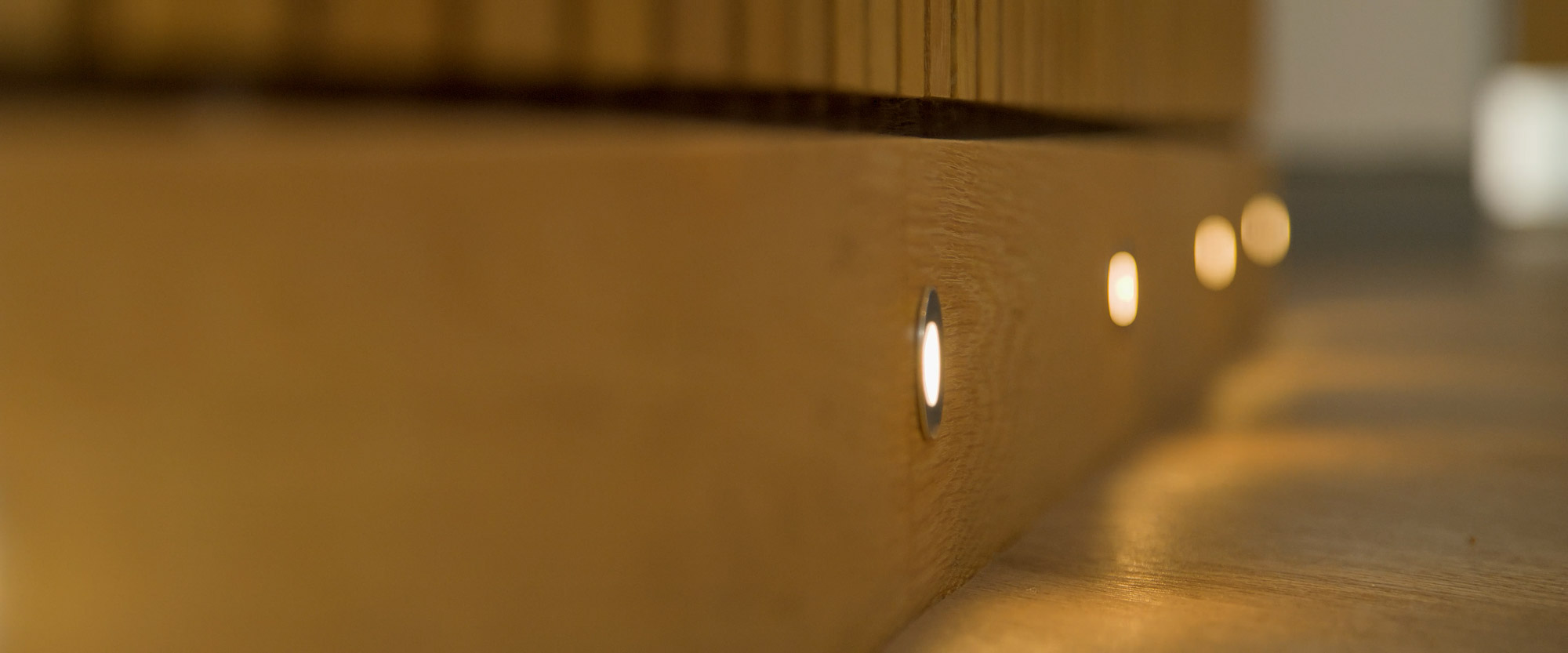 Close-up of small round recessed lights embedded along the side of a wooden surface, casting a warm glow on the floor.