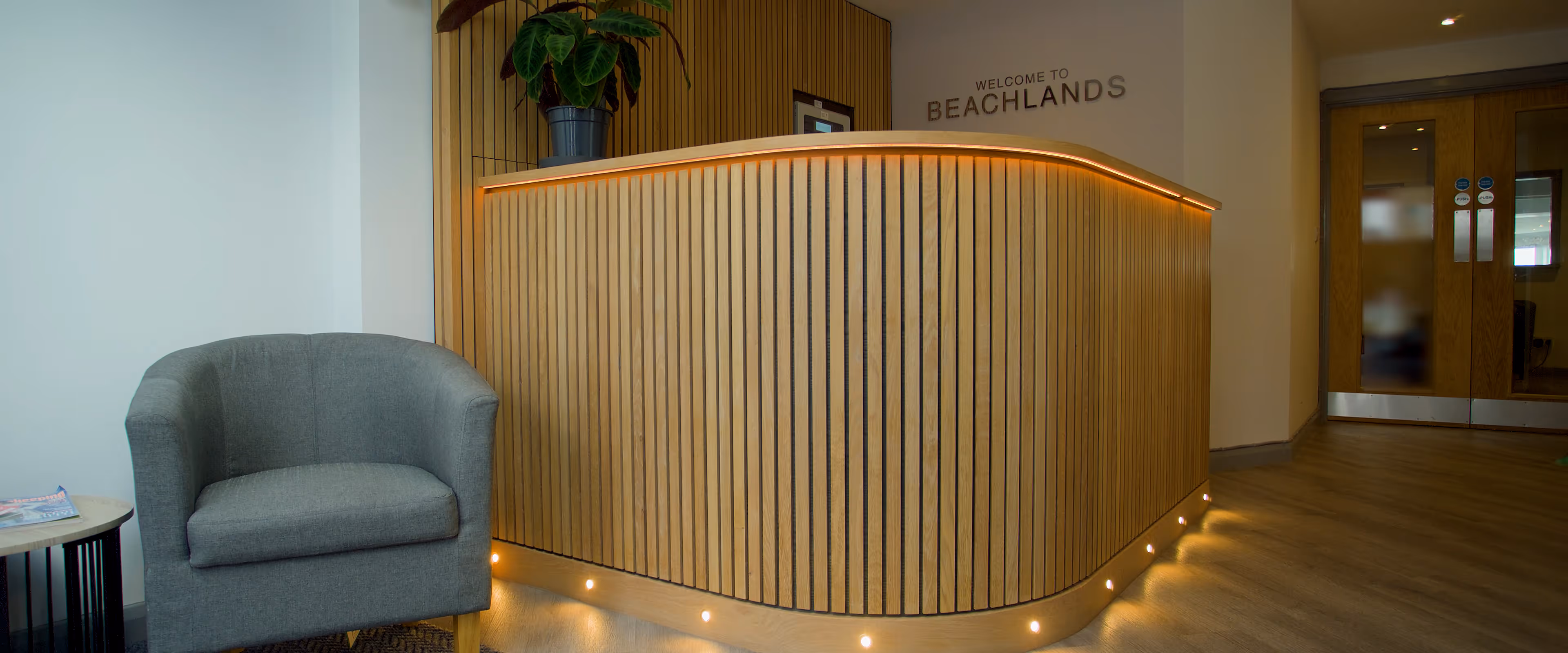 Curved wooden reception desk with warm lighting, gray armchair, potted plant, and double wooden doors in a modern lobby.