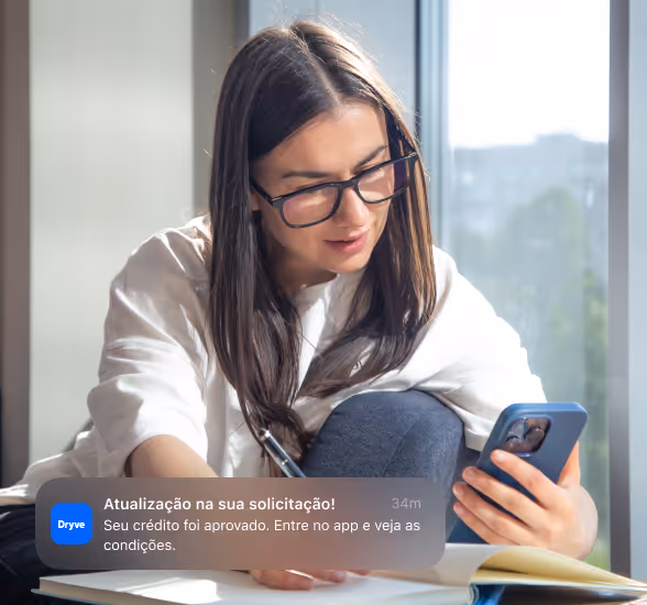 Young woman with glasses looking at her phone while writing in a notebook near a window with city view.