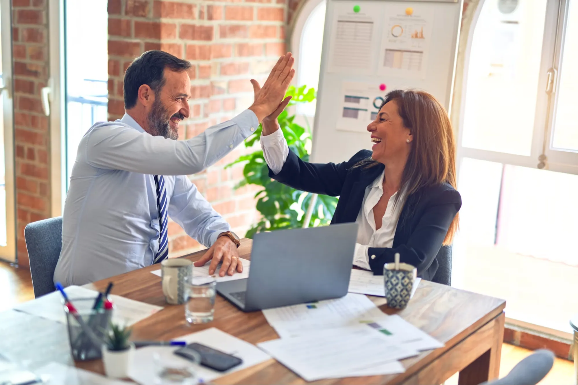 A well-dressed male and female business employee sitting at a wooden table high-five each other. 