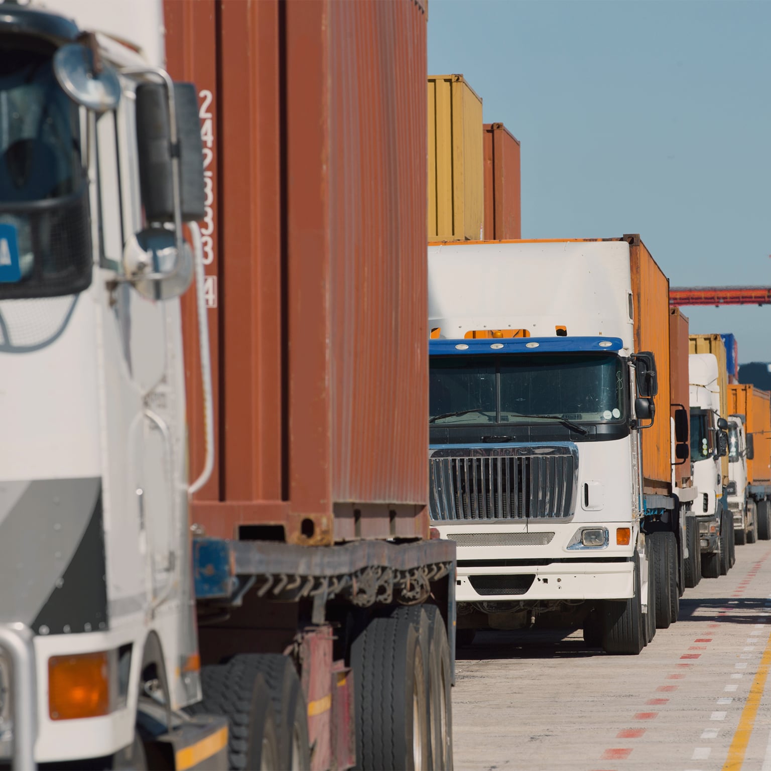 Close-up ground-level view of freight trucks with shipping containers queuing in a line at a cargo terminal.