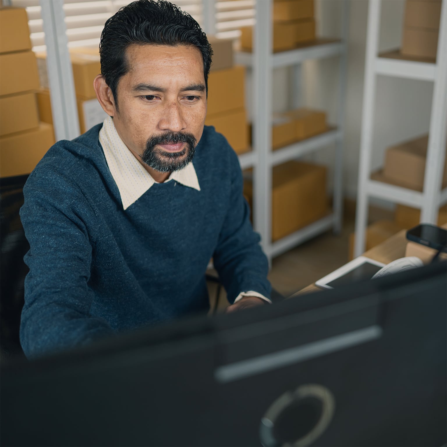 Warehouse manager working at a desktop computer in a storage area with shelves of cardboard boxes in the background.