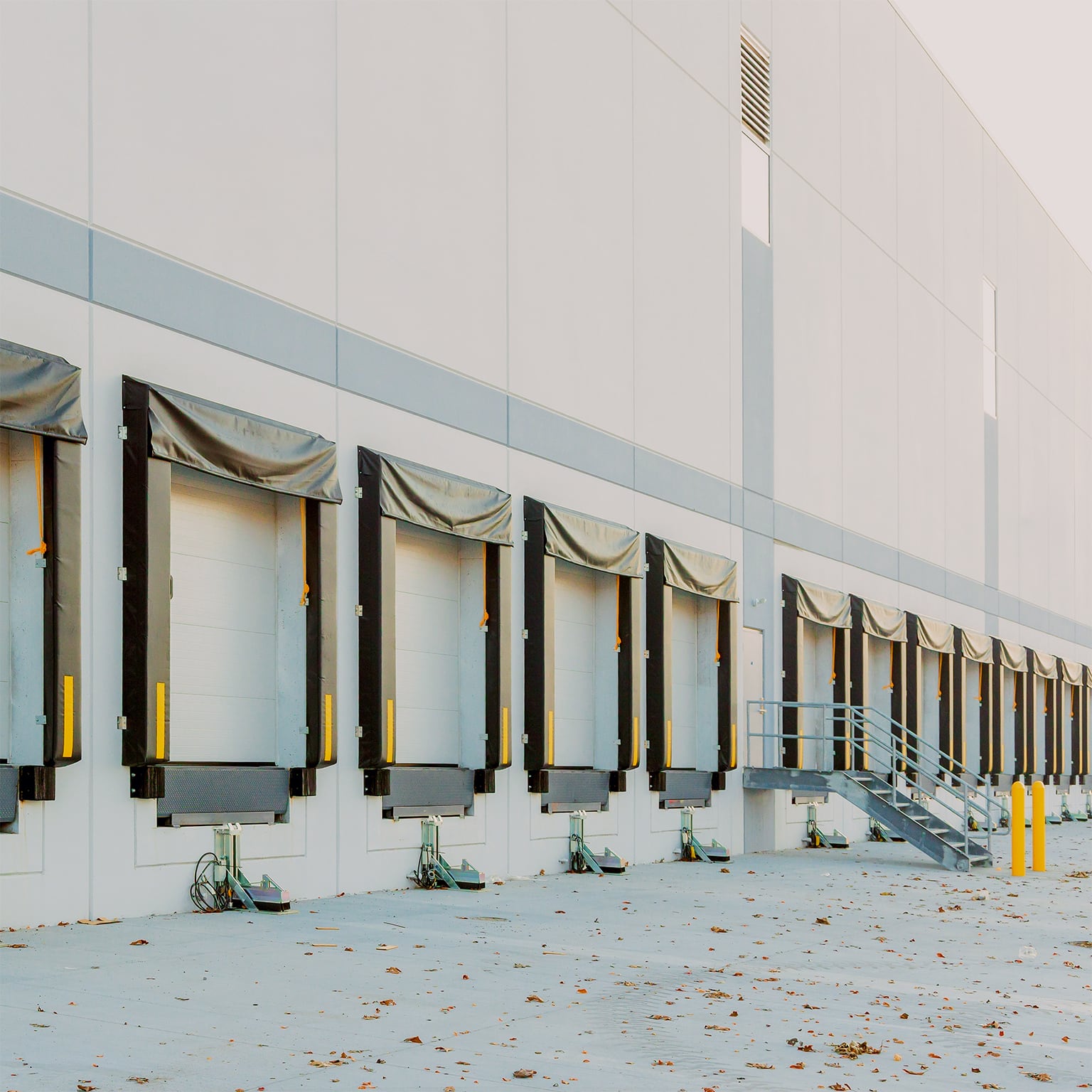 Row of empty loading docks with rubber dock seals along the exterior wall of a modern warehouse, with dock levellers and yellow safety bollards visible.