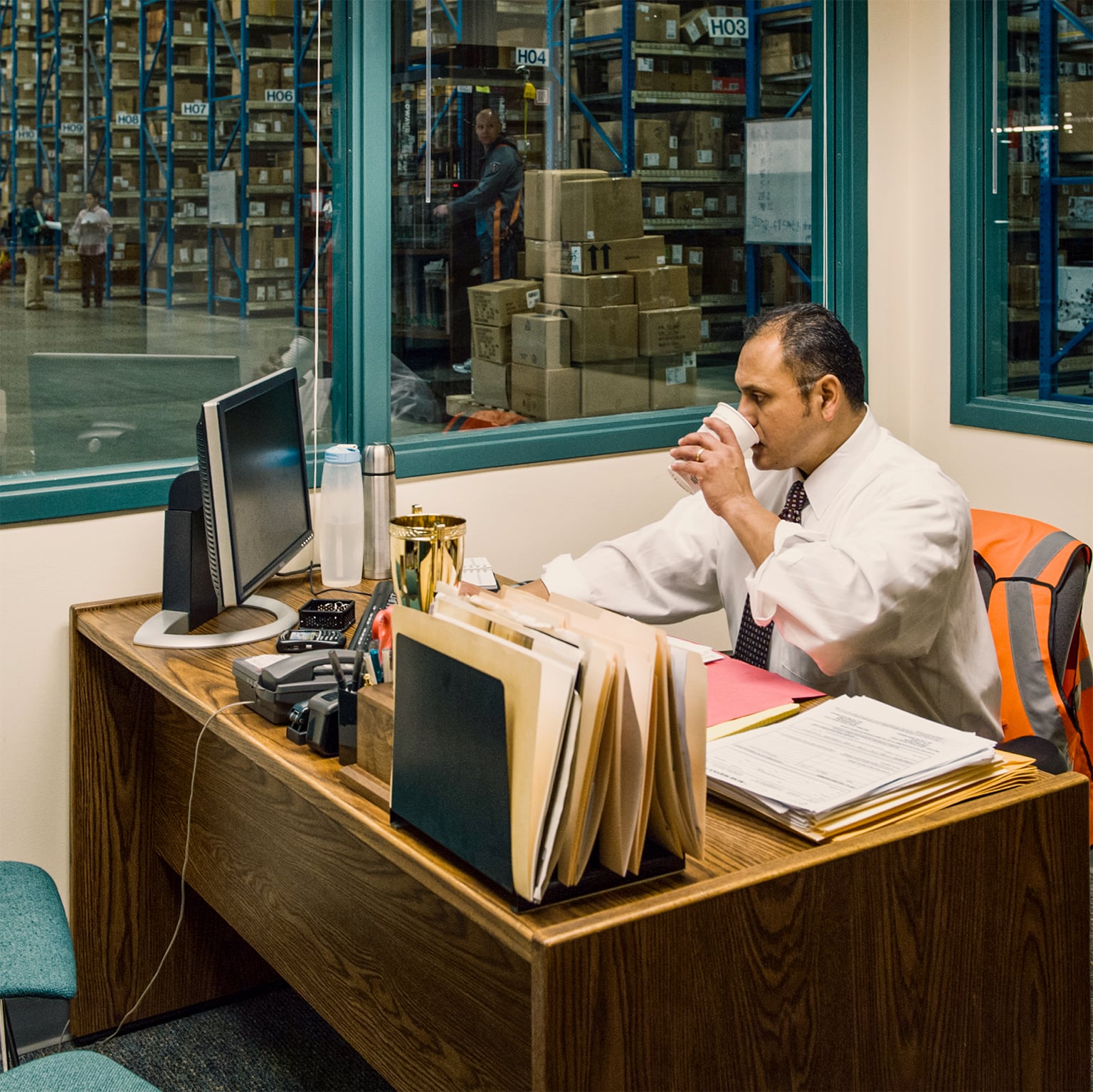 Warehouse manager sipping coffee at his desk in a glass-walled office overlooking the warehouse floor with blue pallet racking and stacked boxes visible through the window.
