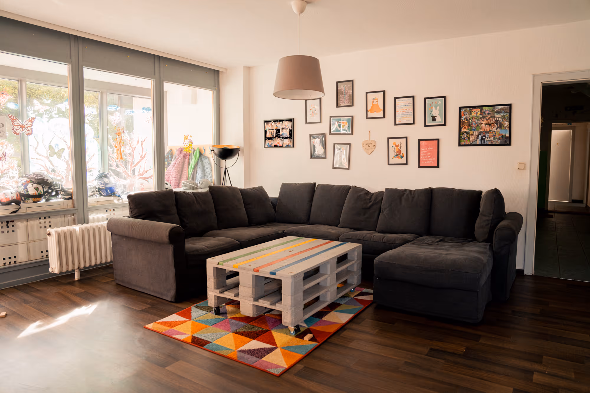 Gemütliches Wohnzimmer mit einem dunkelgrauen Ecksofa, einem bunten geometrischen Teppich und einem Couchtisch aus Paletten auf Rollen unter einer hängenden beigen Lampe.