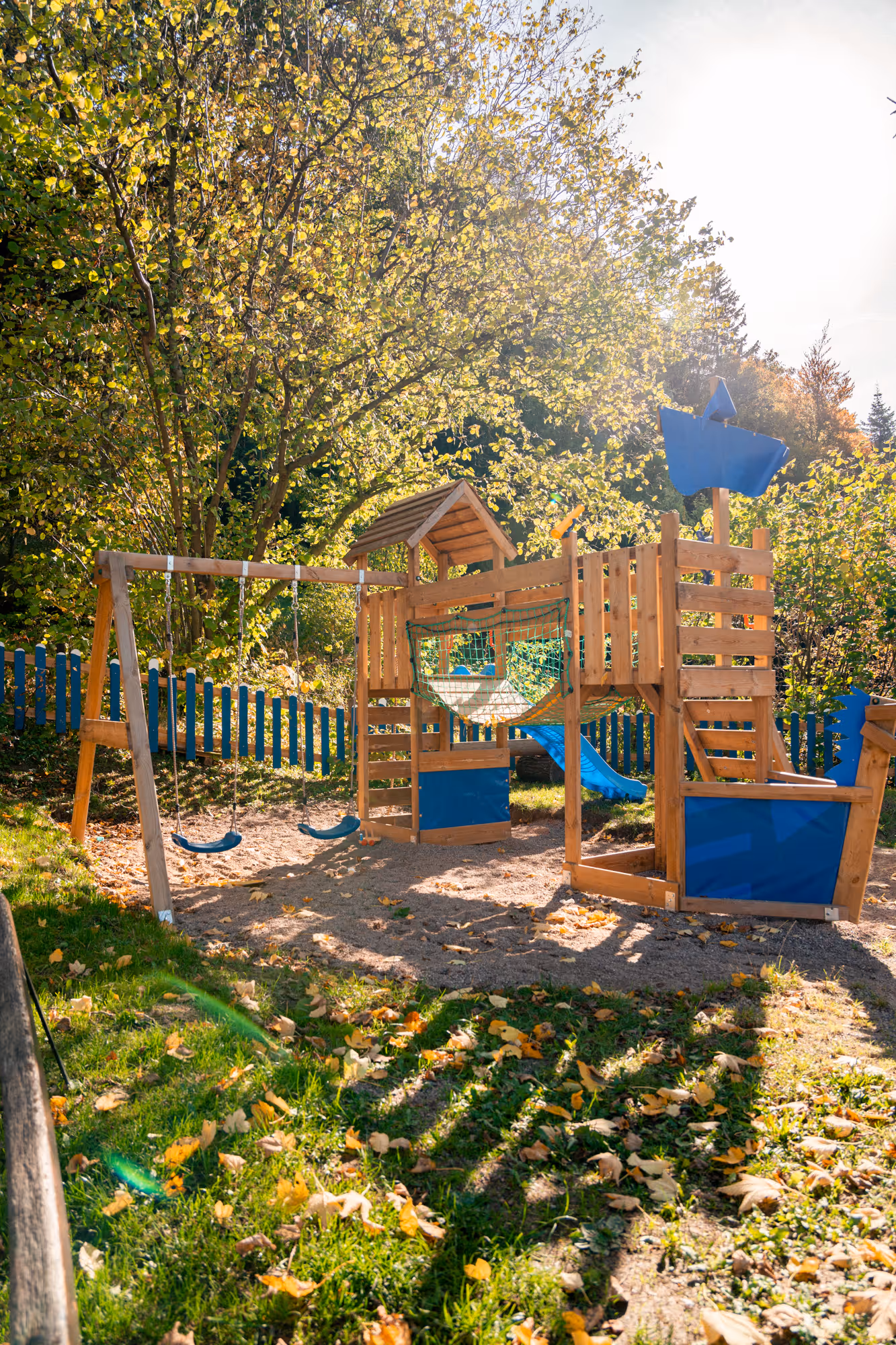 Holzspielplatz mit blauen Akzenten, ausgestattet mit Schaukeln, Rutsche, Kletterleiter und einem Kletternetz, unter Bäumen in einem sonnigen Garten mit blauem Lattenzaun.