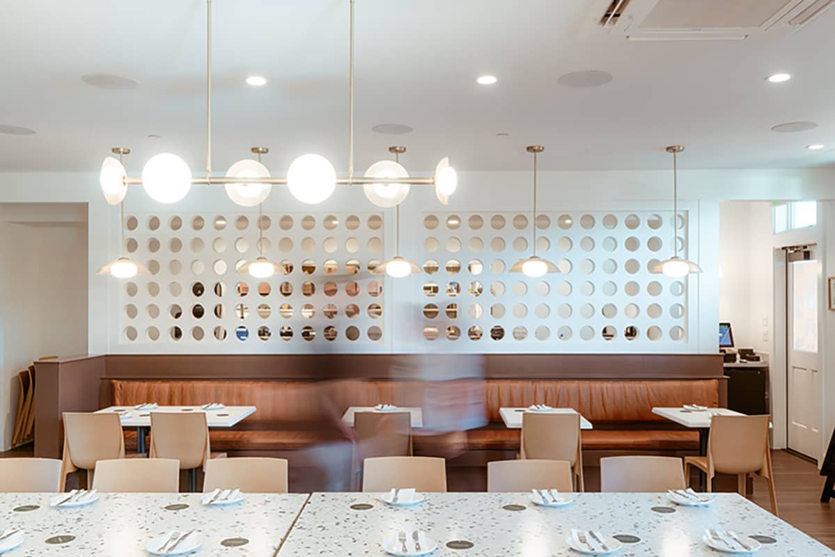Modern restaurant dining area with terrazzo tables, tan chairs, leather bench, and hanging globe lights.
