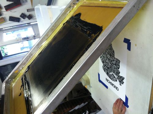 Someone holding up an inked screen printing screen with black ink printing the RVA typographic map on paper below it.