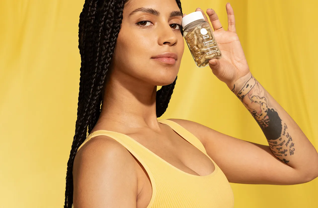 Woman with braided hair wearing yellow holding a bottle of capsules labeled Essential for Women.