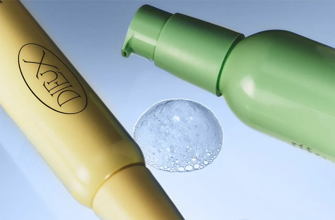 Yellow and green cosmetic bottles next to a transparent foam drop on a light surface.