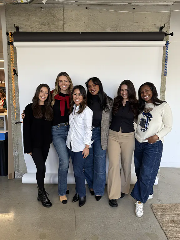 Six diverse women from the Dreamday team smiling and standing in front of a white photo backdrop indoors.