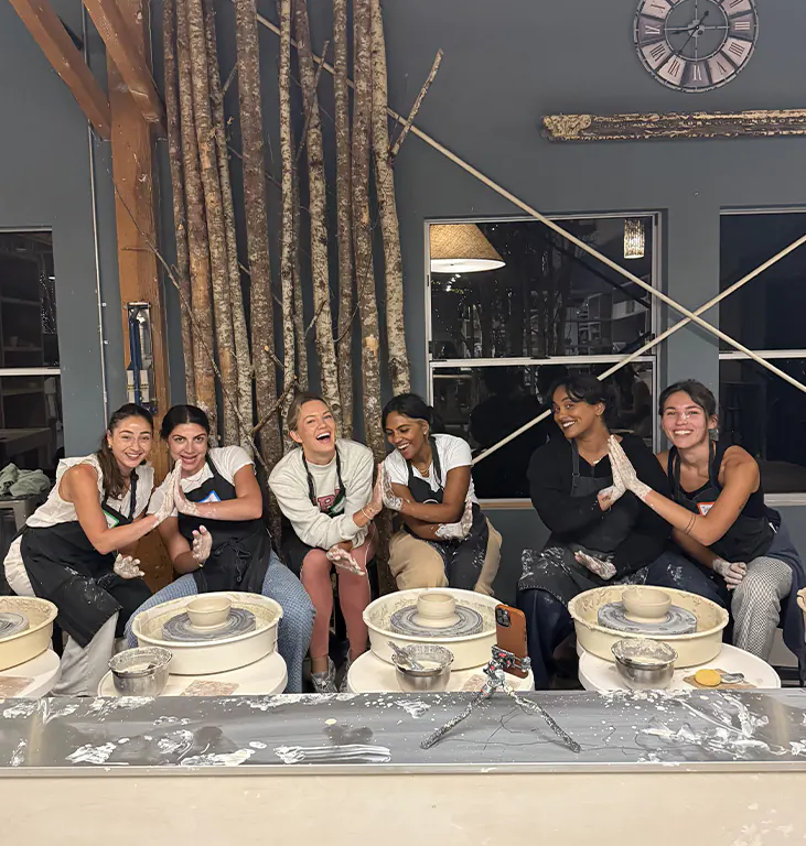 Six women from the Dreamday team smiling and clapping hands in a pottery studio with ceramic wheels and clay bowls.
