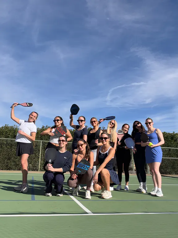 Part of the Dreamday team on a paddle tennis court holding paddles and smiling under a blue sky.