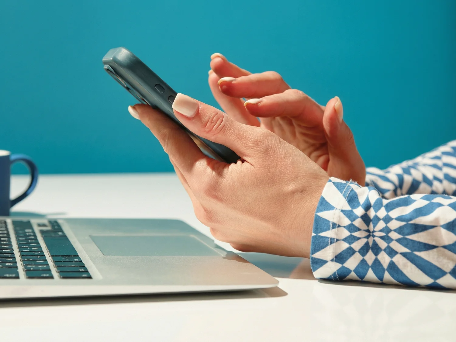 Person holding and using a smartphone beside a laptop on a white desk with a blue mug nearby.