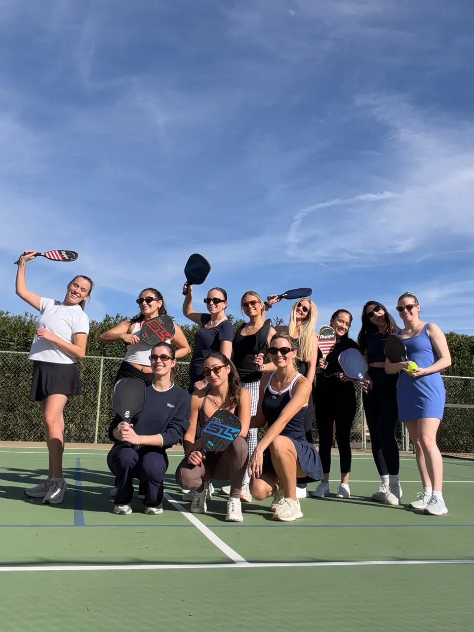 Group of women from the Dreamday team posing with pickleball paddles and ball on an outdoor court under a blue sky.