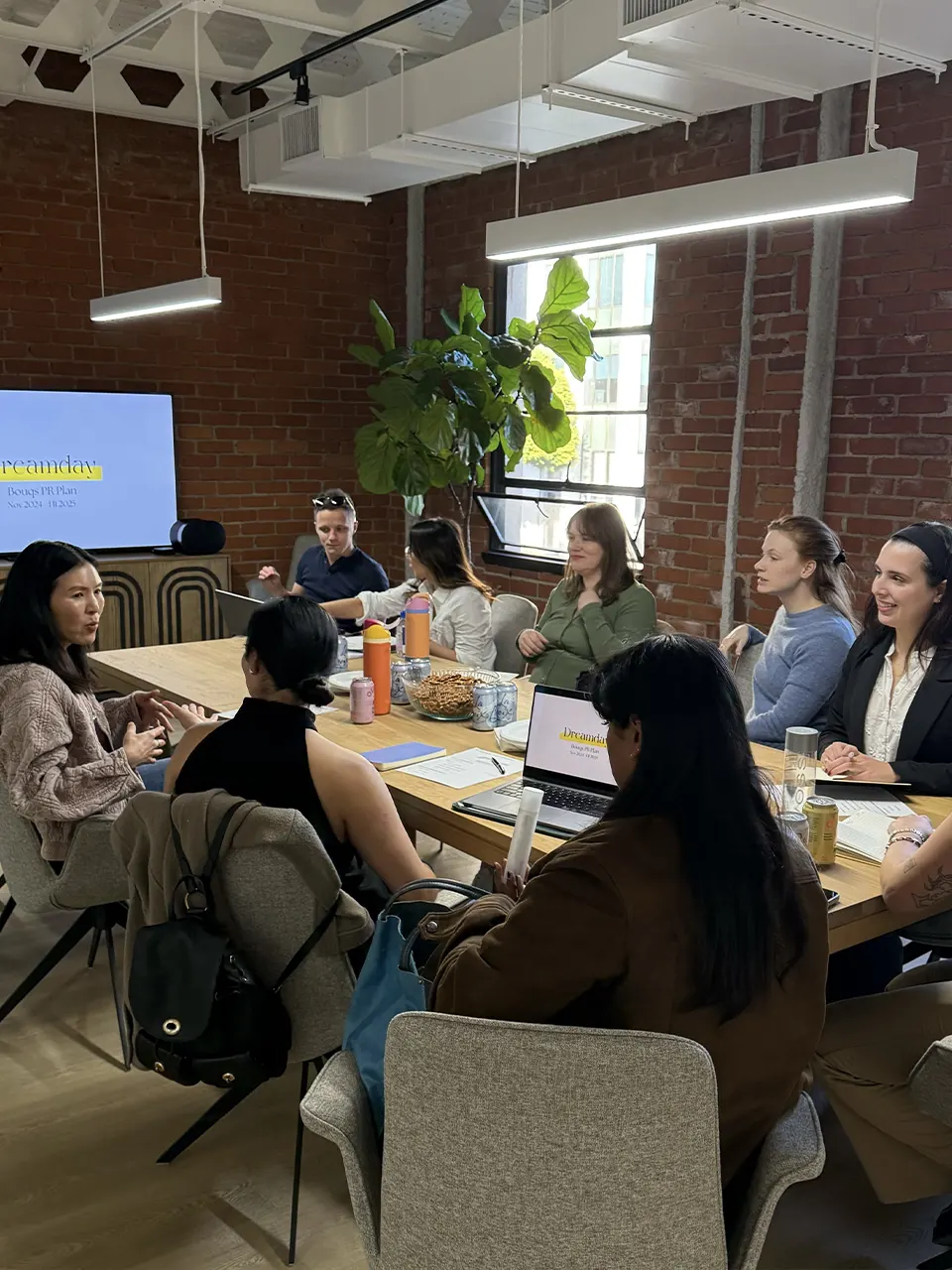 Dreamday team meeting in a modern office with brick walls, discussing plans around a table with laptops and snacks.
