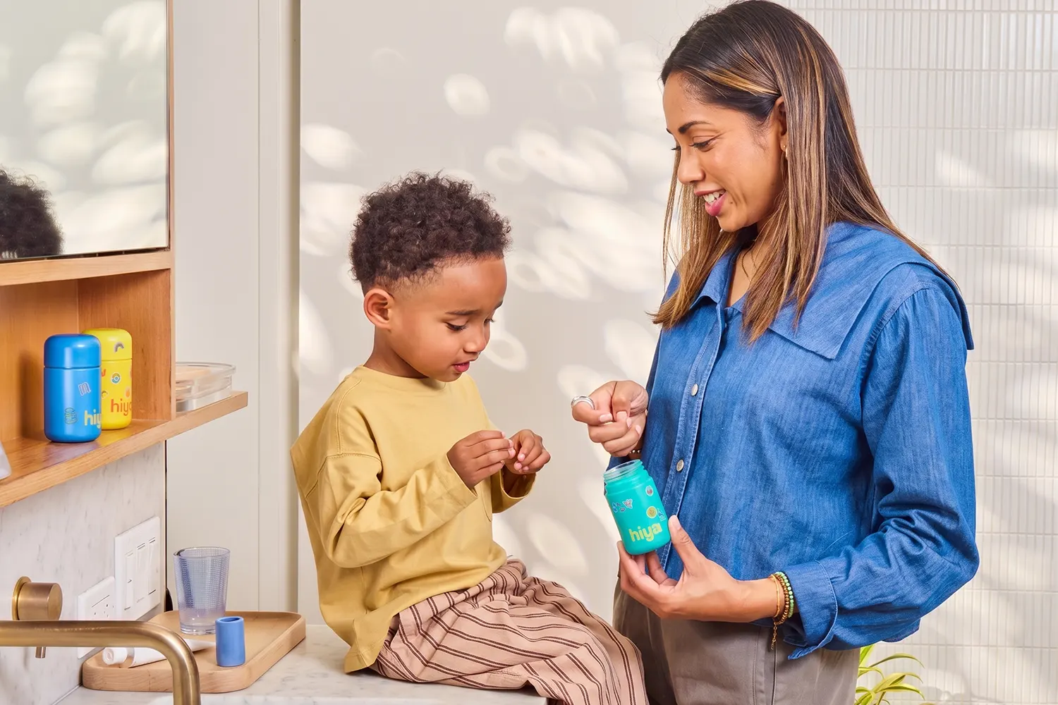 Woman in a blue shirt gives a young boy seated on a counter a small item from a blue Hiya bottle.