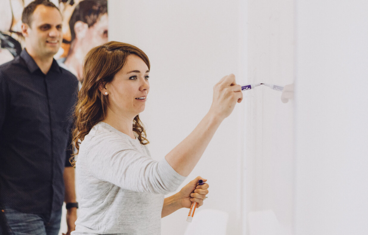 Woman writing on a whiteboard with a man watching in the background.