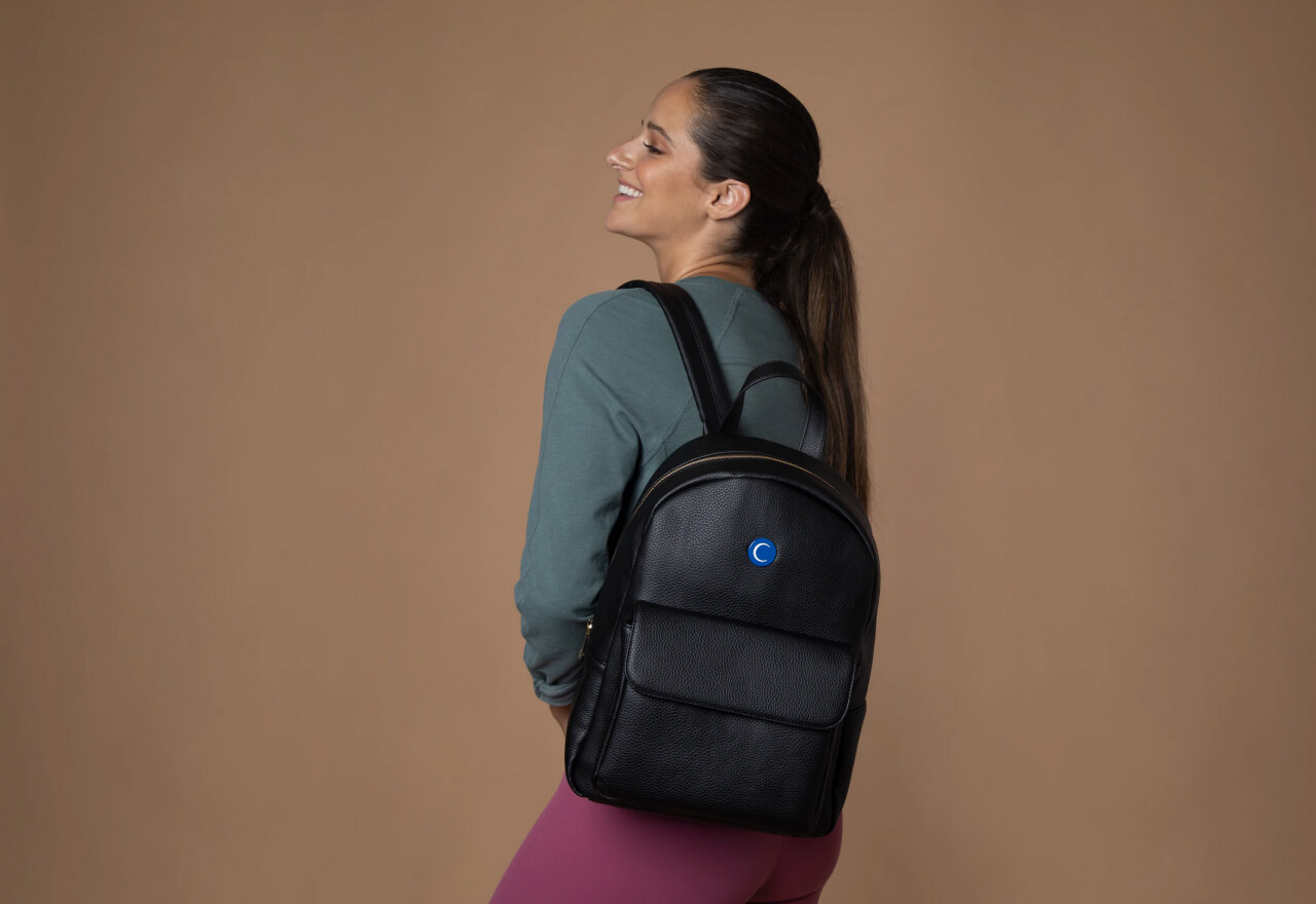 Smiling woman with a ponytail wearing a teal long-sleeve shirt and pink leggings, carrying a black leather backpack on her back against a plain brown background.