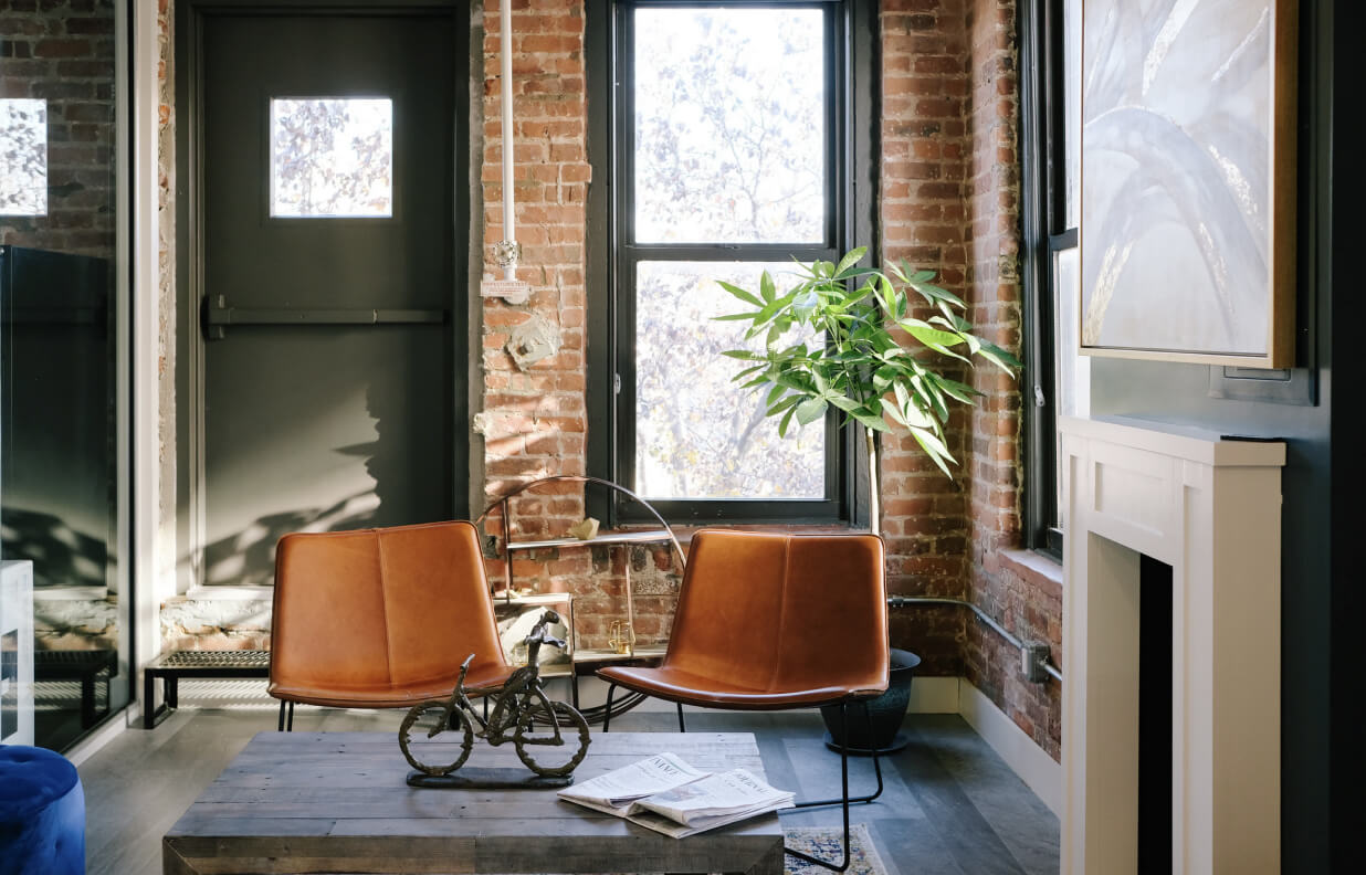 Cozy room with exposed brick walls, two orange leather chairs, a wooden coffee table with a decorative bicycle sculpture and newspapers, and a potted plant by a window.