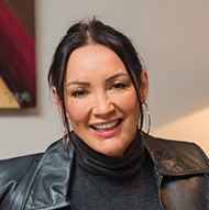 Smiling woman with dark hair wearing a black turtleneck and leather jacket against a neutral background.