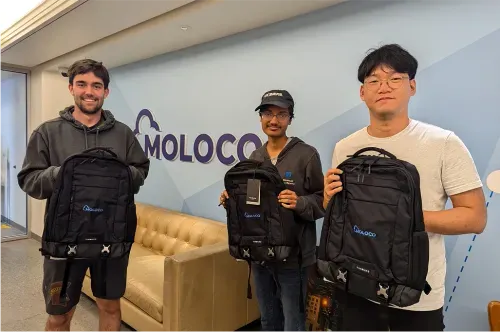 Three Moloco interns in a modern office setting, each holding a black backpack with a 'Moloco' logo, stand in front of a wall with the company name.