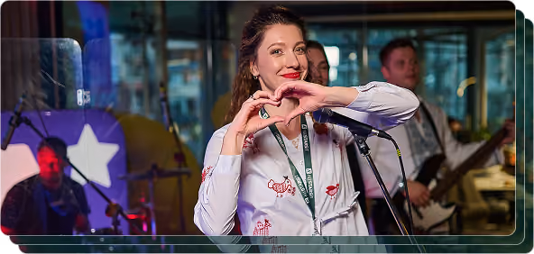 Woman making a heart shape with hands near a microphone, band playing in background indoors.
