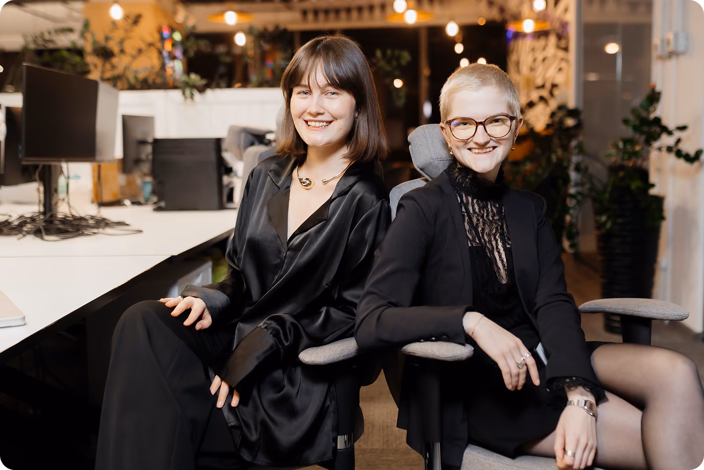 Two women in black outfits sit smiling on office chairs in a modern workspace with computers nearby.