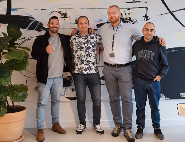 Four men standing indoors side by side, smiling, with a large plant and abstract wall art behind them.