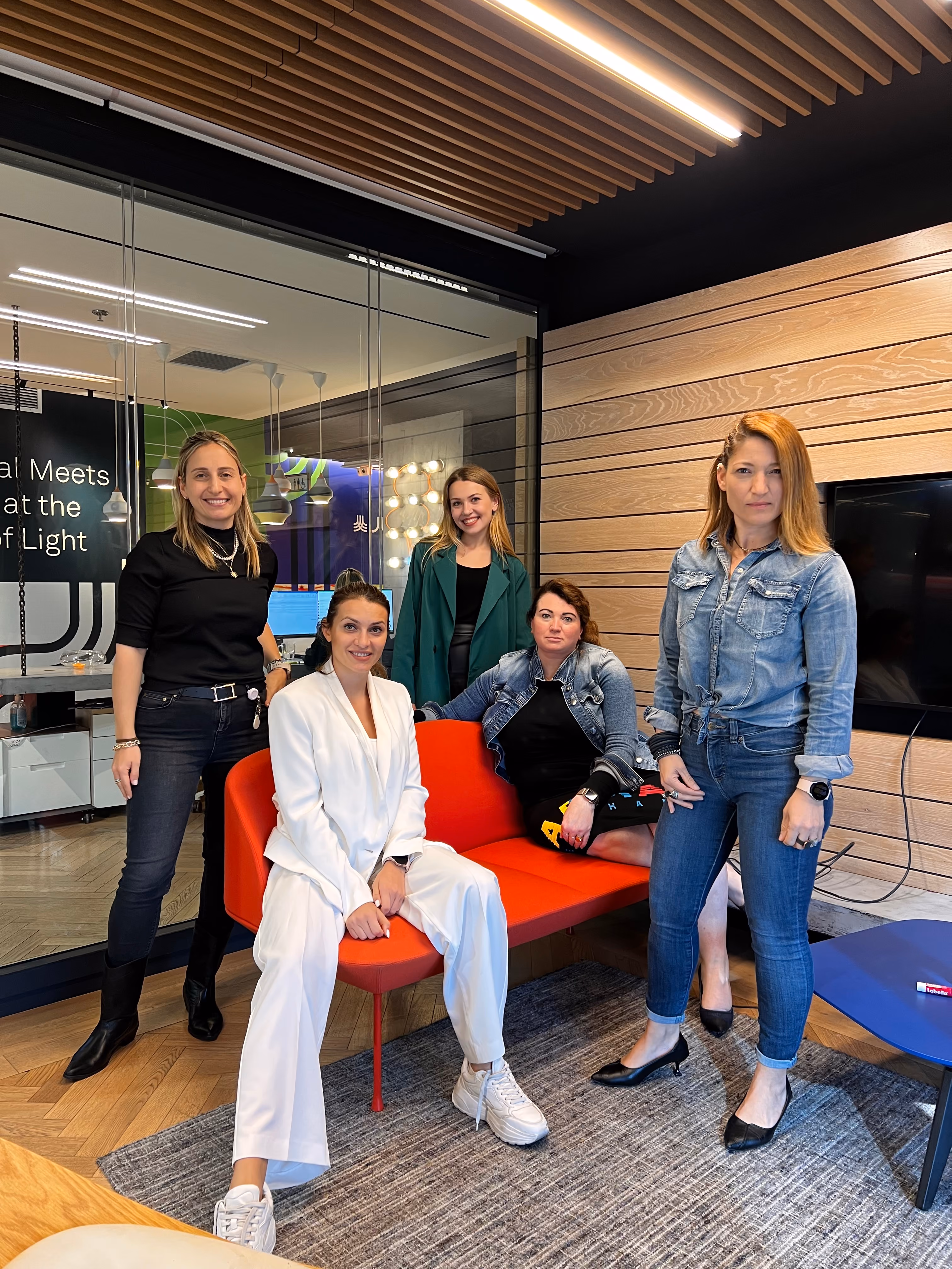 Five women posing in an office lounge area with a red couch and wood-paneled wall.
